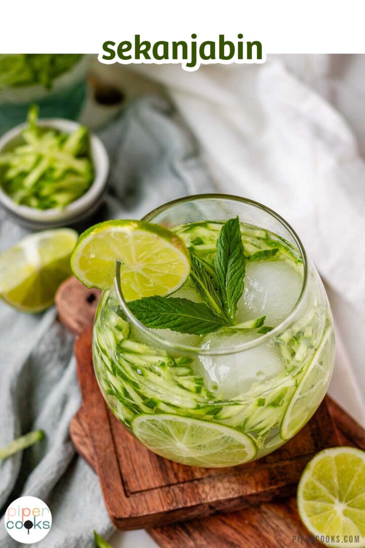 A drink in a glass garnished with julienned cucumber, lime slices, and a sprig of mint with the text sekajabin.