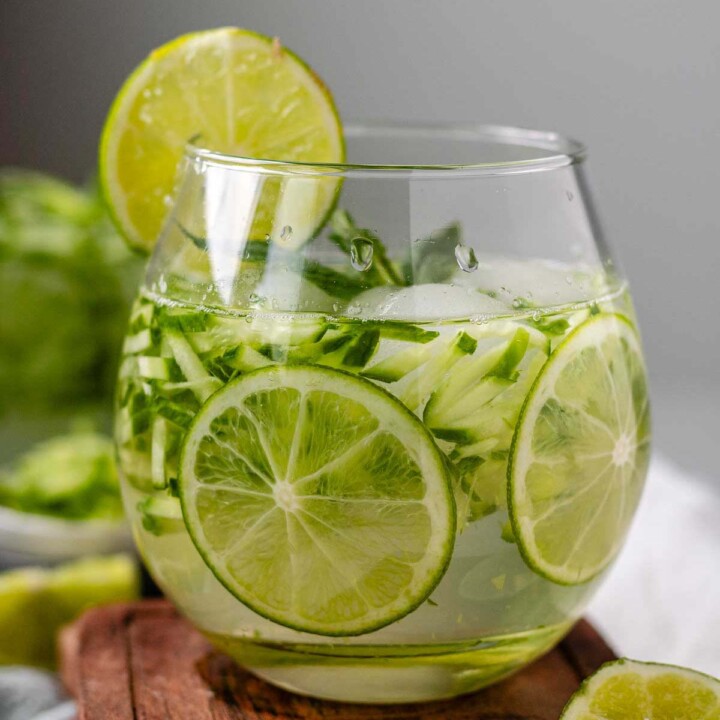 A drink in a glass garnished with julienned cucumber, lime slices, and a sprig of mint.