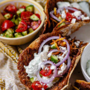 Tacos filled with meat, tomato, cucumber, red onion, feta, white sauce, and shoestring fries with the text Greek-Inspired Smash Tacos.