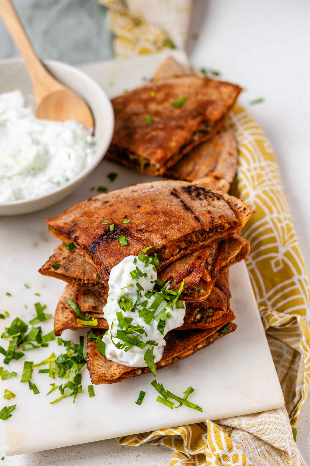 A stack of meat-filled fried pitas with tzatziki sauce and fresh parsley on top.