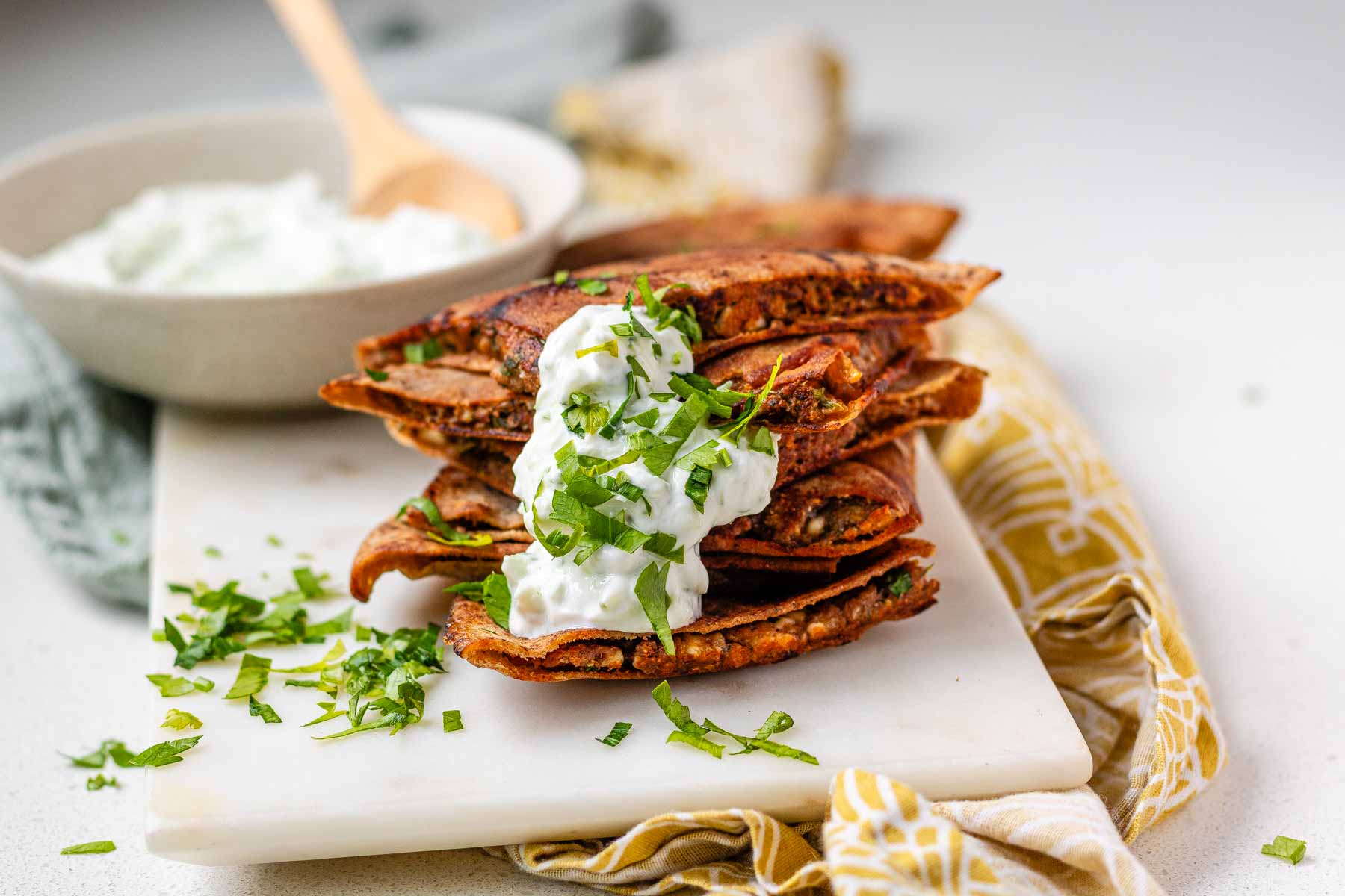A stack of meat-filled fried pitas with tzatziki sauce and fresh parsley on top.