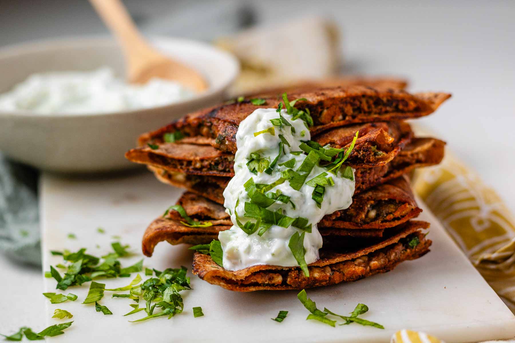 A stack of meat-filled fried pitas with tzatziki sauce and fresh parsley on top.