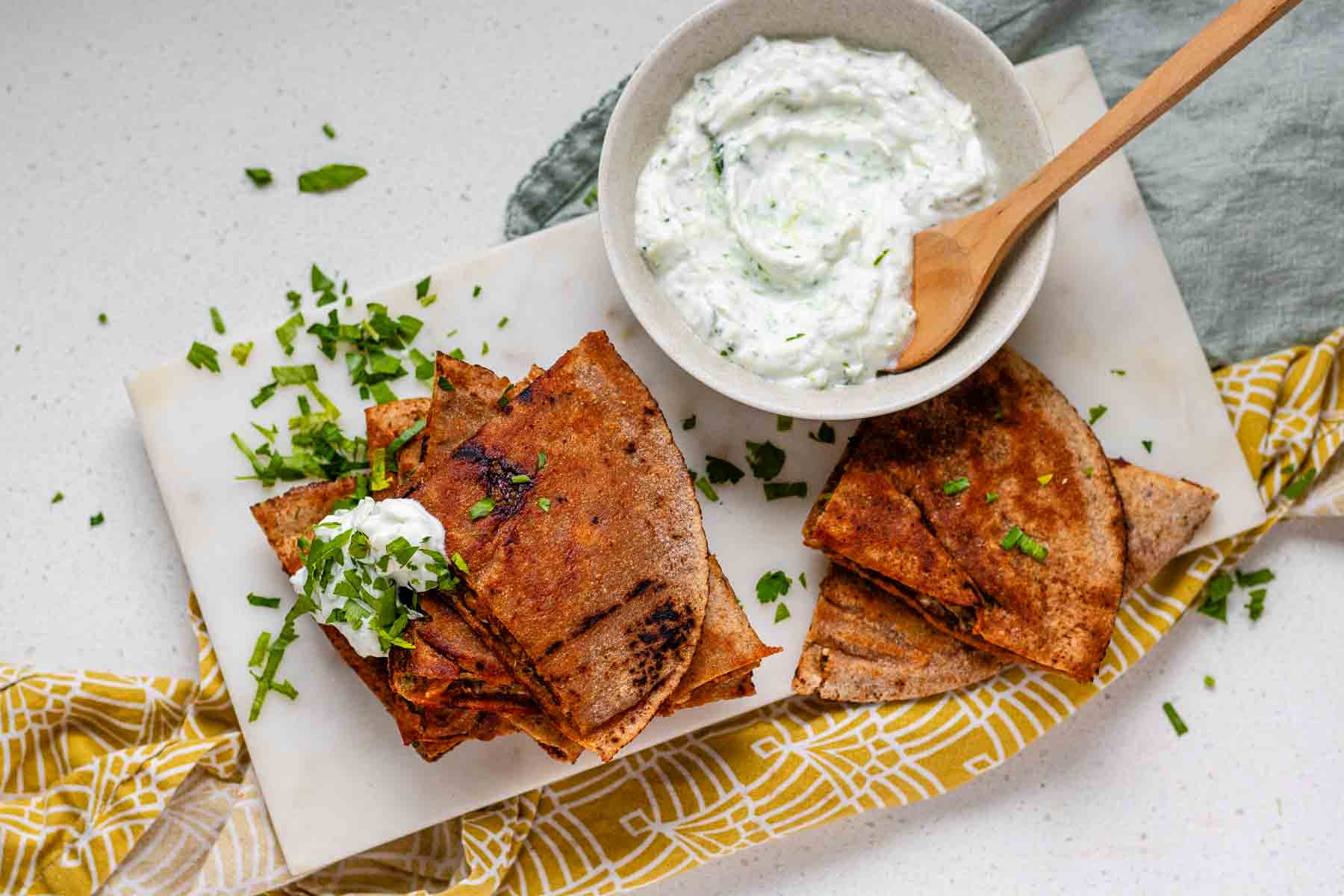 A stack of meat-filled fried pitas with tzatziki sauce and fresh parsley on top.