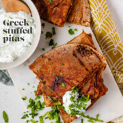 A stack of meat-filled fried pitas with tzatziki sauce and fresh parsley on top, with the text Greek stuffed pitas.