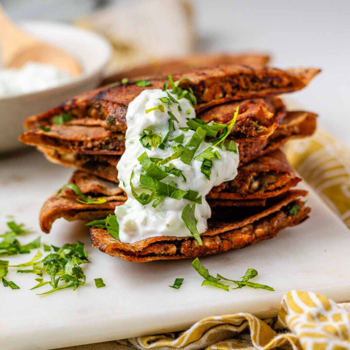 A stack of meat-filled fried pitas with tzatziki sauce and fresh parsley on top.