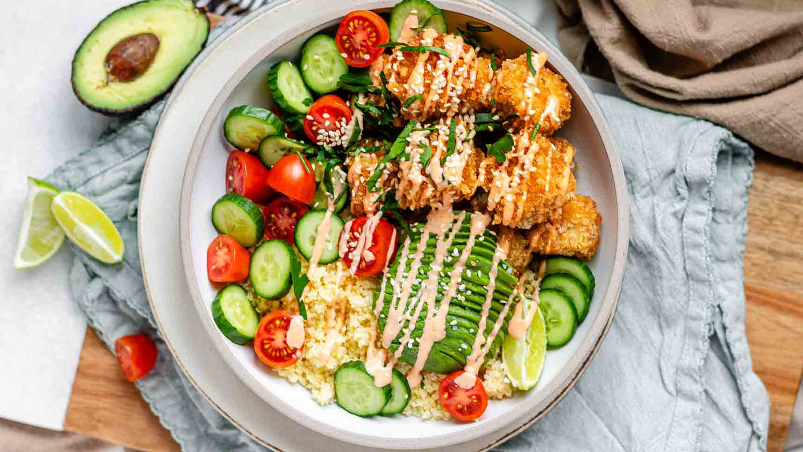 A bowl full of couscous, cucumber, tomato, avocado, and breaded tofu drizzled in sauce.