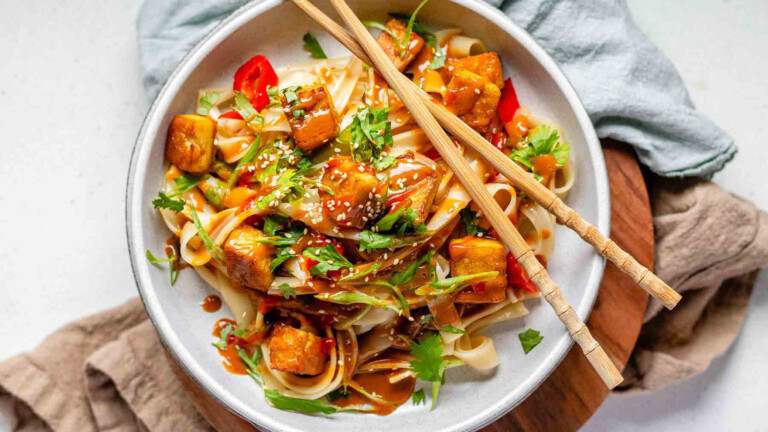 Spicy Peanut Tofu Bowl with rice noodles, red peppers, and creamy peanut sauce with chopsticks.