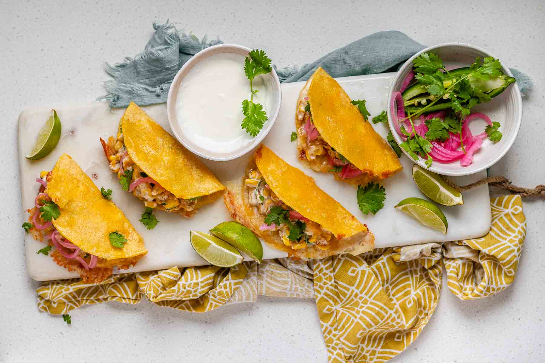 Cheesy crispy corn tacos on a white platter with garlic lime crema and pickled onions and lime wedges.