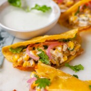 Cheesy crispy corn tacos on a white platter with garlic lime crema and pickled onions and lime wedges.