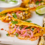 Cheesy crispy corn tacos on a white platter with garlic lime crema and pickled onions and lime wedges.