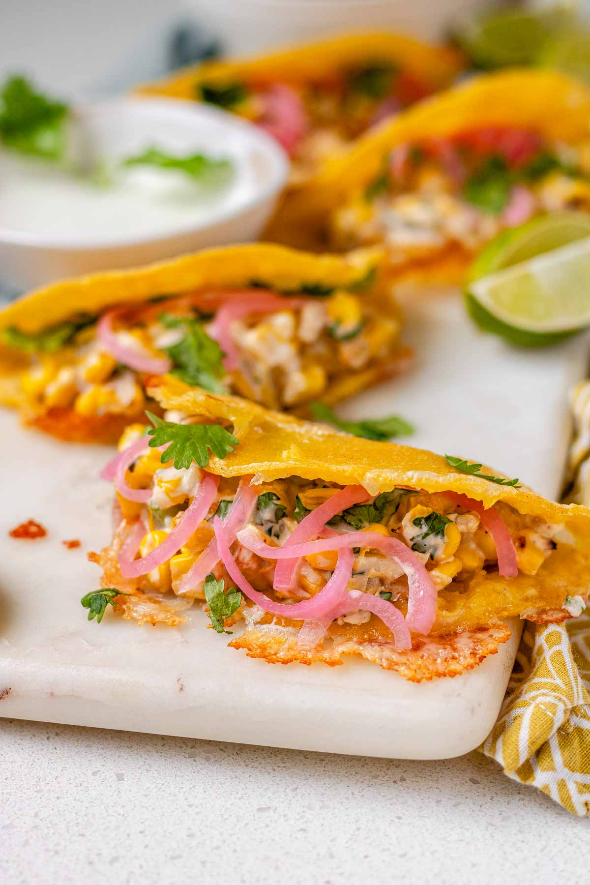 Cheesy crispy corn tacos on a white platter with garlic lime crema and pickled onions and lime wedges.