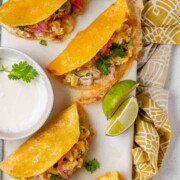 Cheesy crispy corn tacos on a white platter with garlic lime crema and pickled onions and lime wedges.