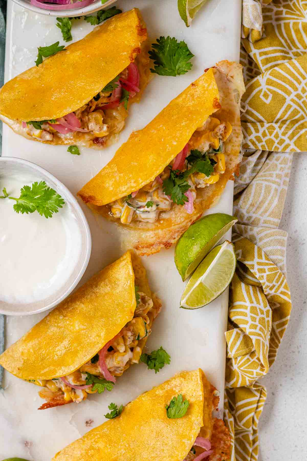 Cheesy crispy corn tacos on a white platter with garlic lime crema and pickled onions and lime wedges.