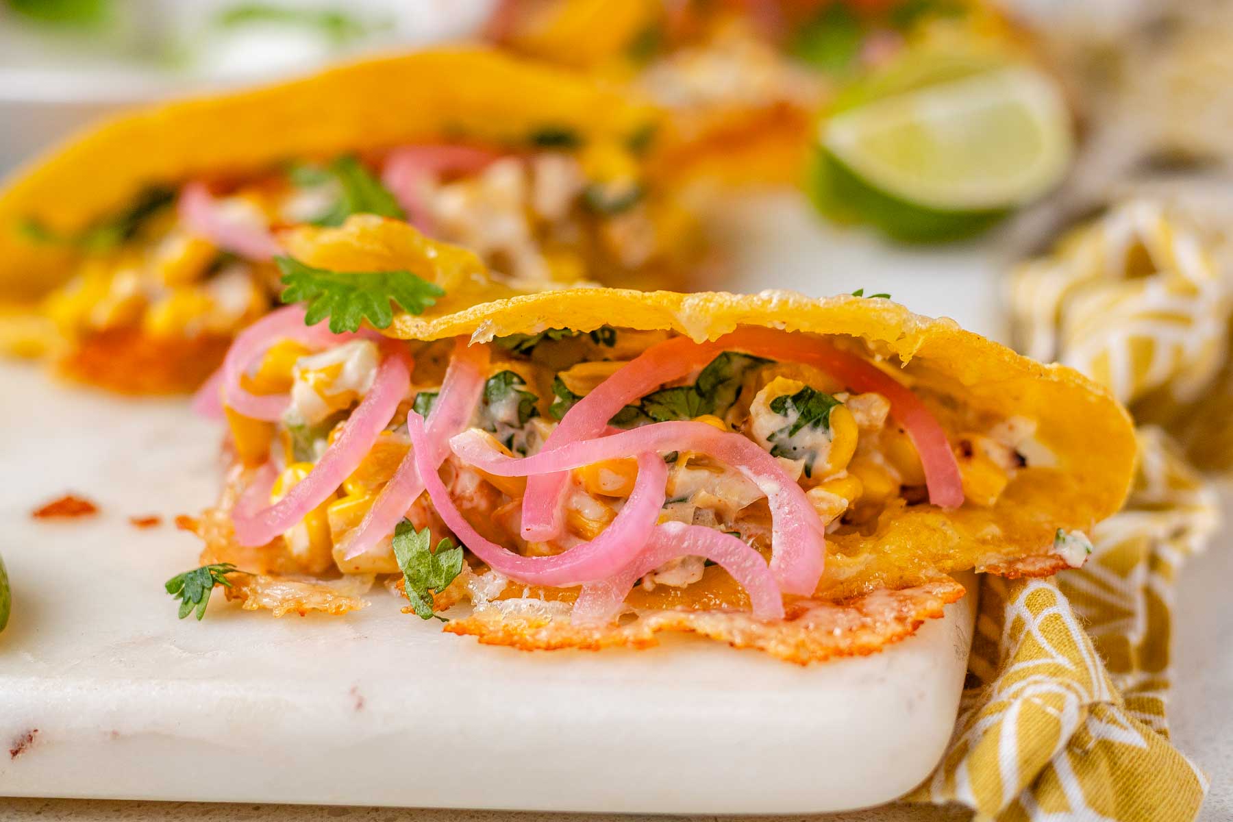Cheesy crispy corn tacos on a white platter with garlic lime crema and pickled onions and lime wedges.
