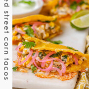 Cheesy crispy corn tacos on a white platter with garlic lime crema and pickled onions and lime wedges with the text baked street corn tacos.