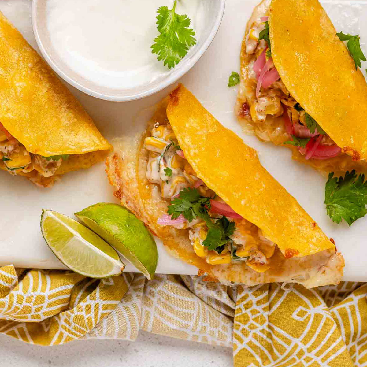 Cheesy crispy corn tacos on a white platter with garlic lime crema and pickled onions and lime wedges.