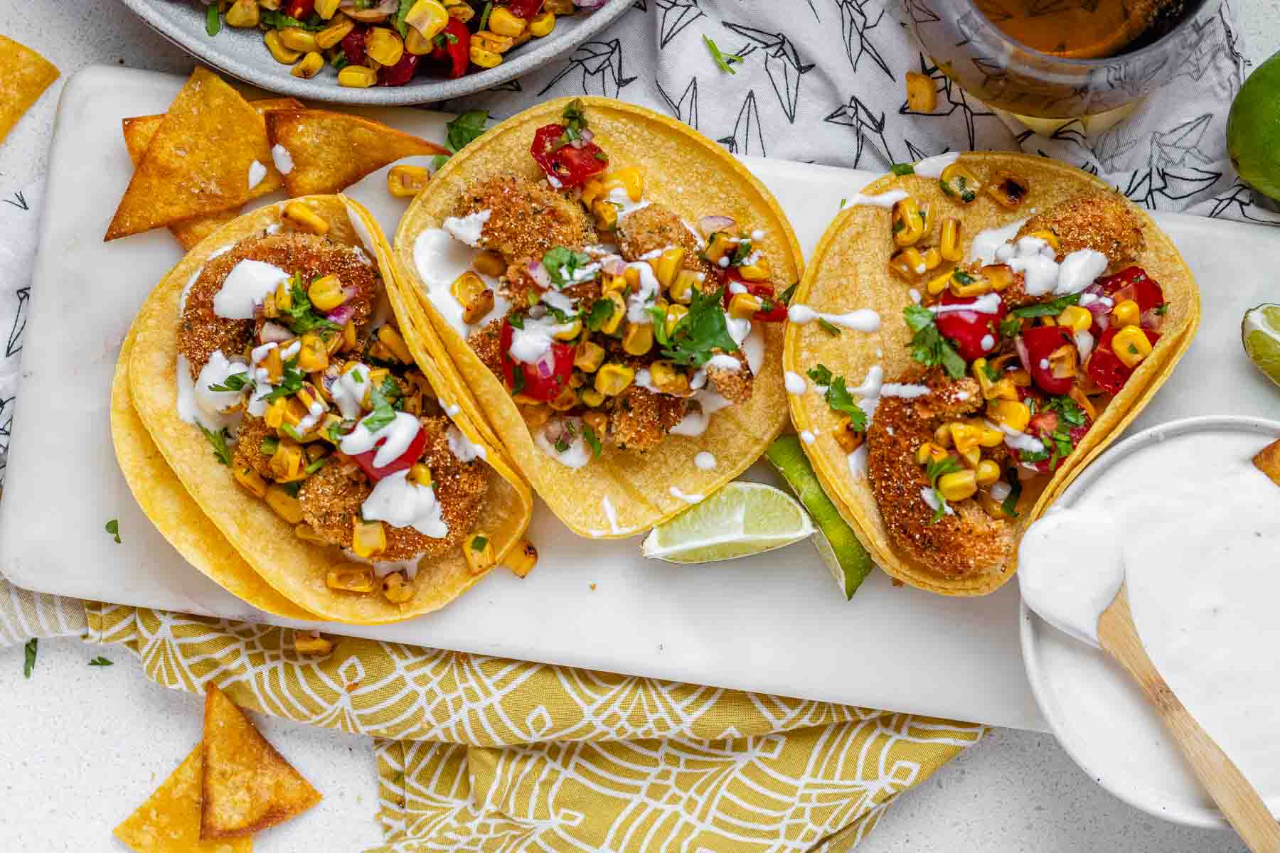 Tacos filled with shrimp and a corn and tomato salsa, on a white platter with garlic lime crema and lime wedges.
