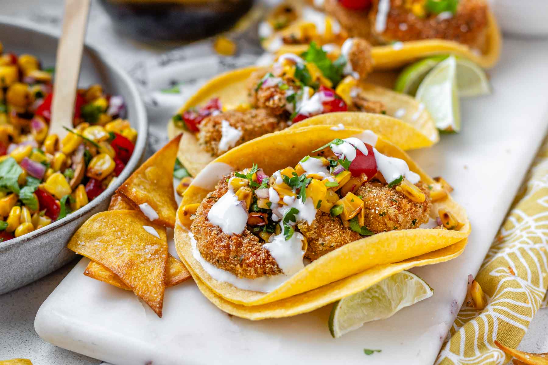 Tacos filled with shrimp and a corn and tomato salsa, on a white platter with garlic lime crema and lime wedges.