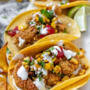 Tacos filled with shrimp and a corn and tomato salsa, on a white platter with garlic lime crema and lime wedges.