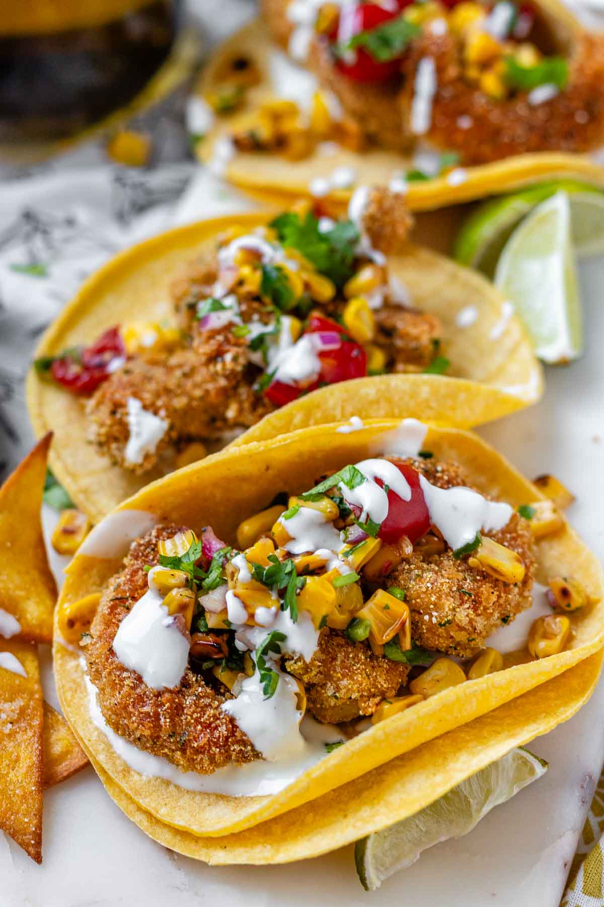 Tacos filled with shrimp and a corn and tomato salsa, on a white platter with garlic lime crema and lime wedges.