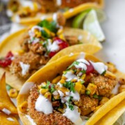 Tacos filled with shrimp and a corn and tomato salsa, on a white platter with garlic lime crema and lime wedges.