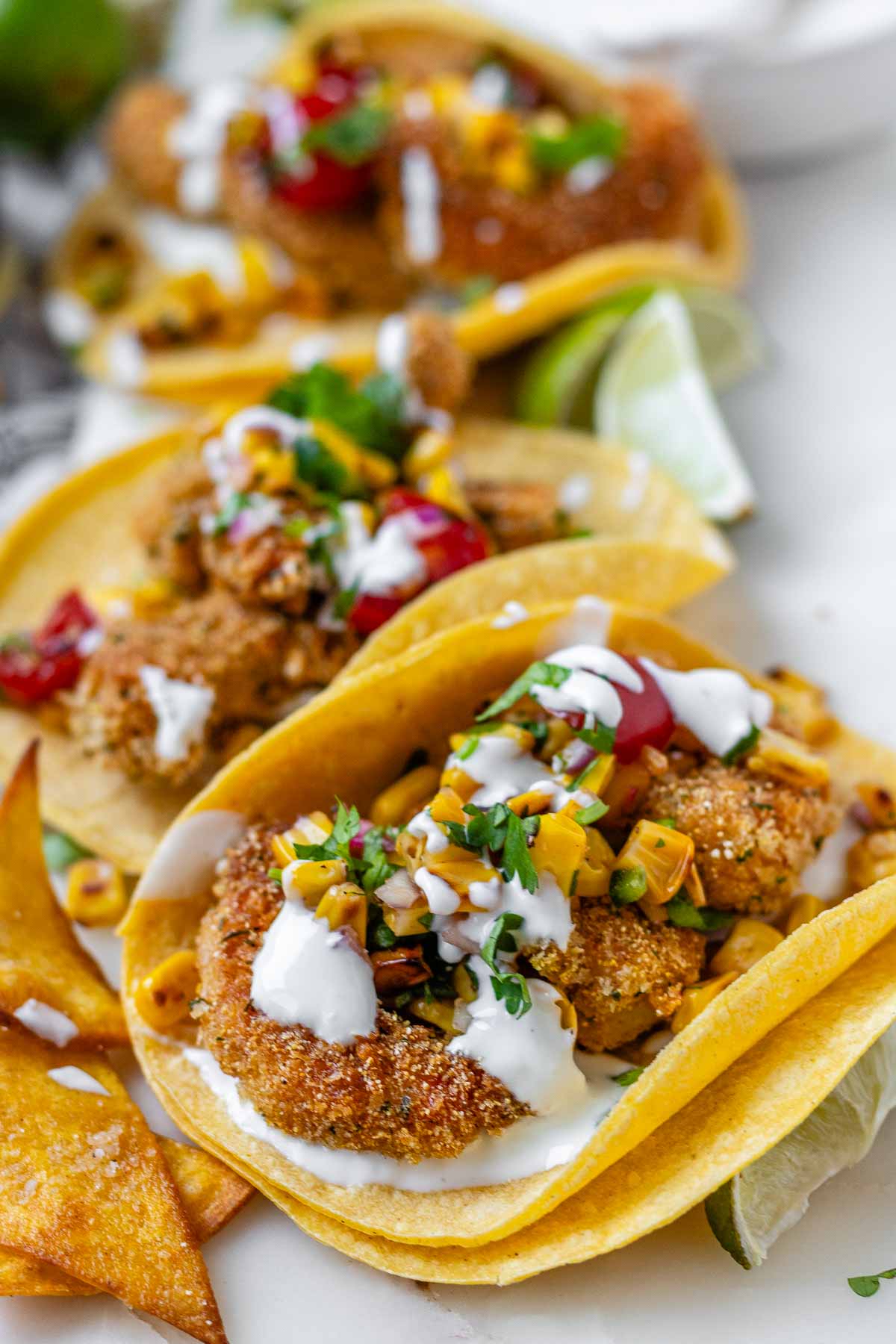 Tacos filled with shrimp and a corn and tomato salsa, on a white platter with garlic lime crema and lime wedges.