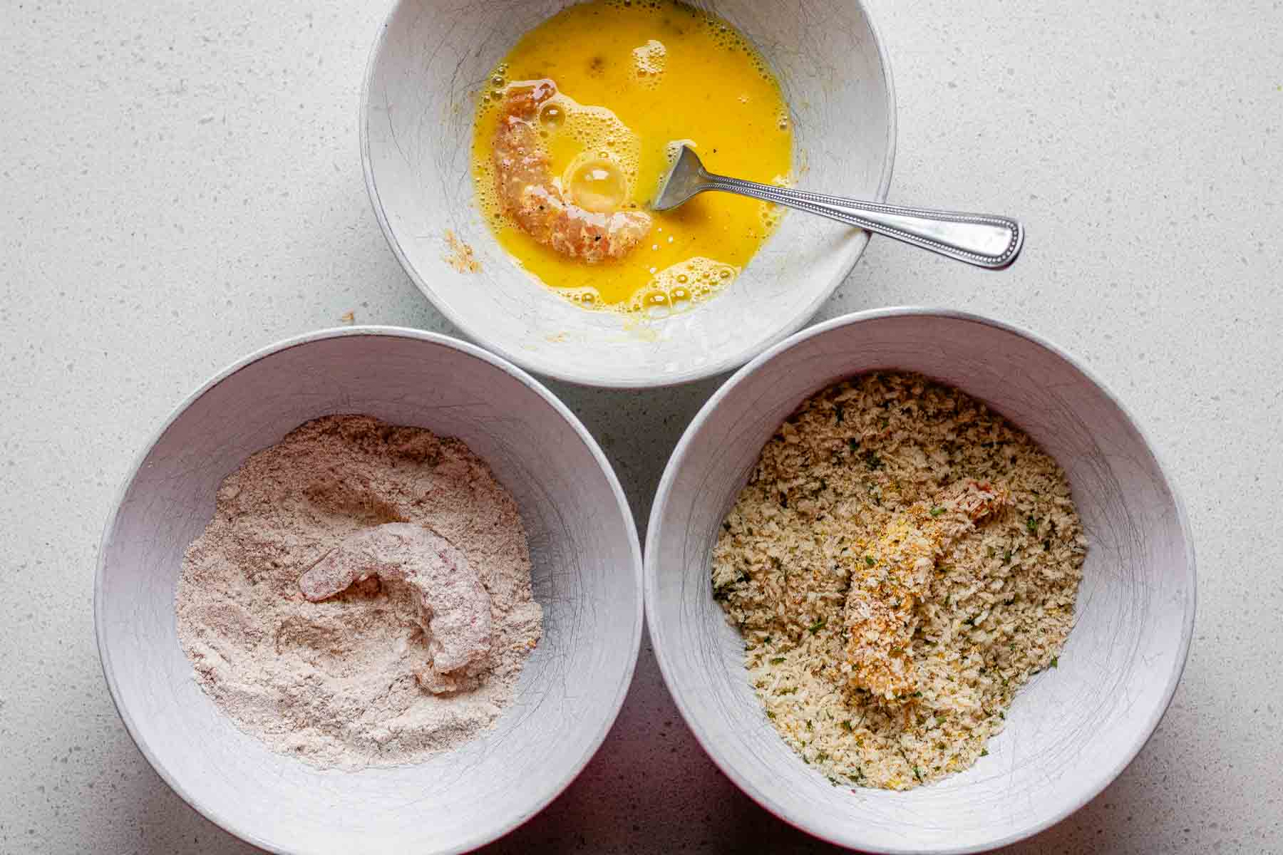 Three bowls each with one shrimp, coated in flour, egg, and breadcrumbs, one ingredient per bowl.