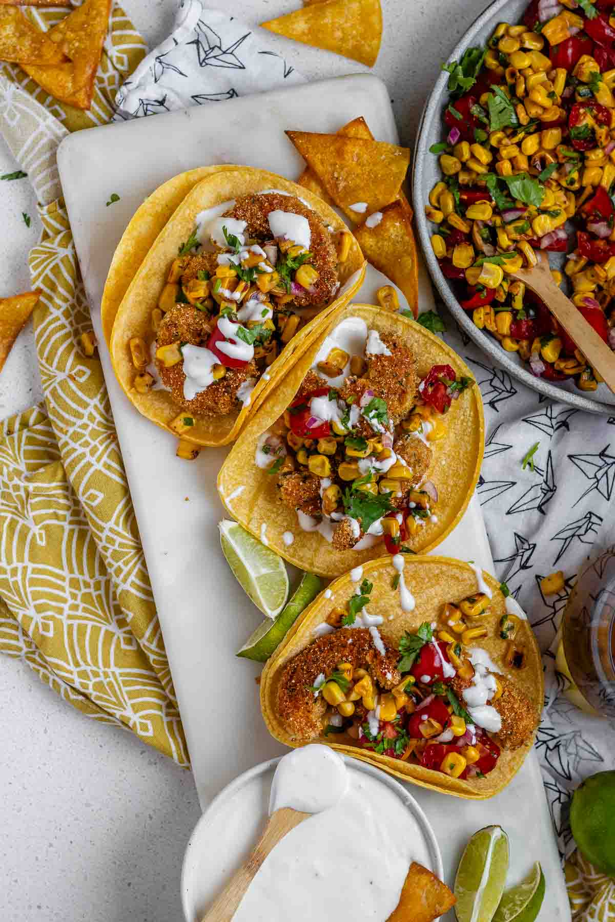 Tacos filled with shrimp and a corn and tomato salsa, on a white platter with garlic lime crema and lime wedges.
