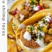 Tacos filled with shrimp and a corn and tomato salsa, on a white platter with garlic lime crema and lime wedges with the text crispy shrimp tacos.