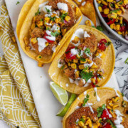 Tacos filled with shrimp and a corn and tomato salsa, on a white platter with garlic lime crema and lime wedges with the text crispy shrimp tacos.