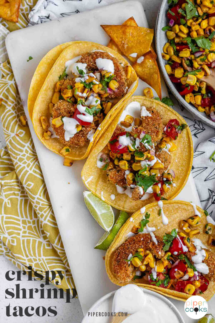 Tacos filled with shrimp and a corn and tomato salsa, on a white platter with garlic lime crema and lime wedges with the text crispy shrimp tacos.