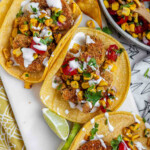 Tacos filled with shrimp and a corn and tomato salsa, on a white platter with garlic lime crema and lime wedges.