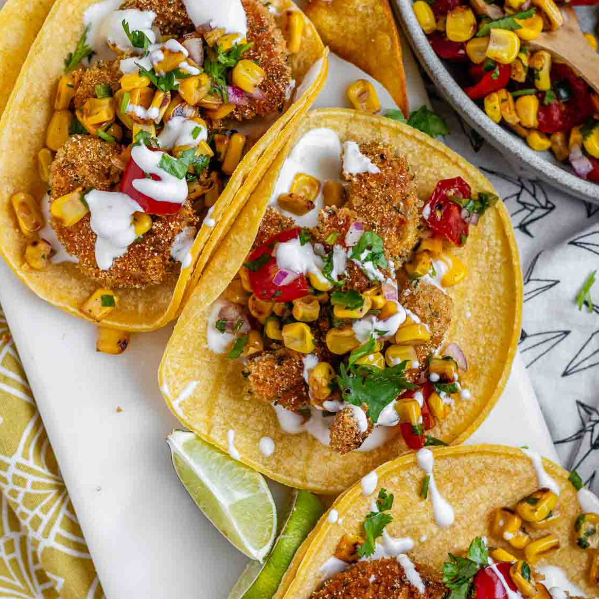 Tacos filled with shrimp and a corn and tomato salsa, on a white platter with garlic lime crema and lime wedges.