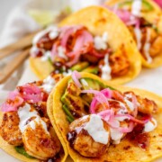 Hot honey Cajun shrimp tacos on a white platter with garlic lime crema and pickled onions.