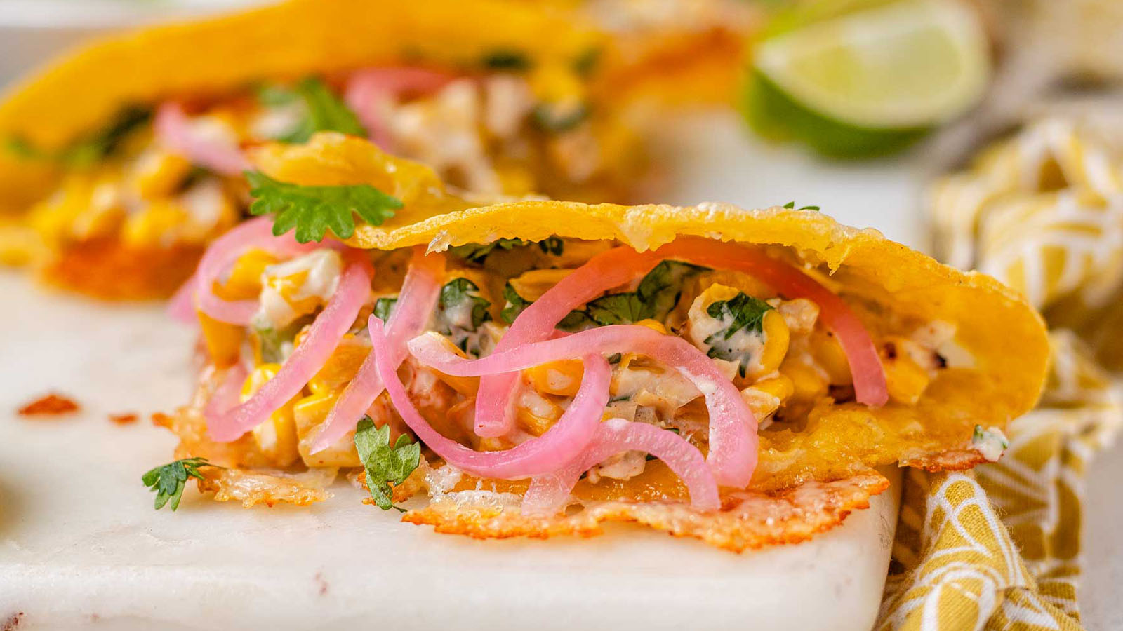 Cheesy crispy corn tacos on a white platter with garlic lime crema and pickled onions and lime wedges.