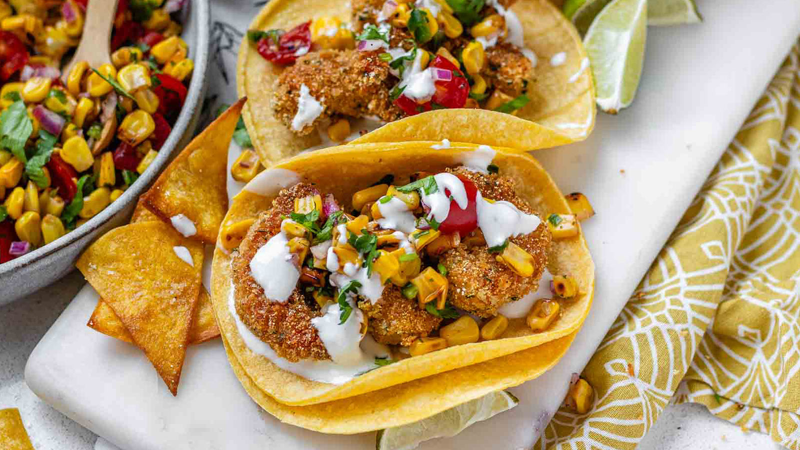 Tacos filled with shrimp and a corn and tomato salsa, on a white platter with garlic lime crema and lime wedges.
