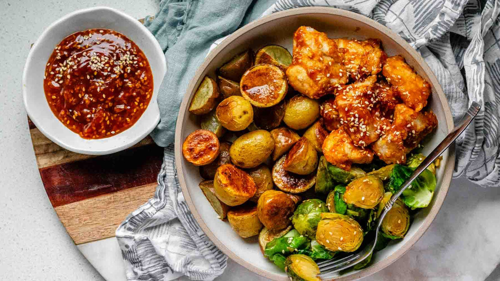 Plated serving of spicy sesame chicken, baby potatoes, and Brussels sprouts with extra sesame sauce on the side.