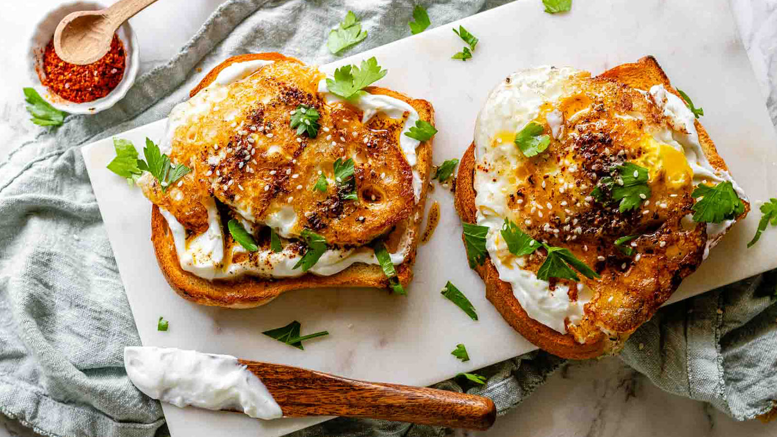 Two slices of toast on a white board topped with white spread, fried eggs, and cilantro.