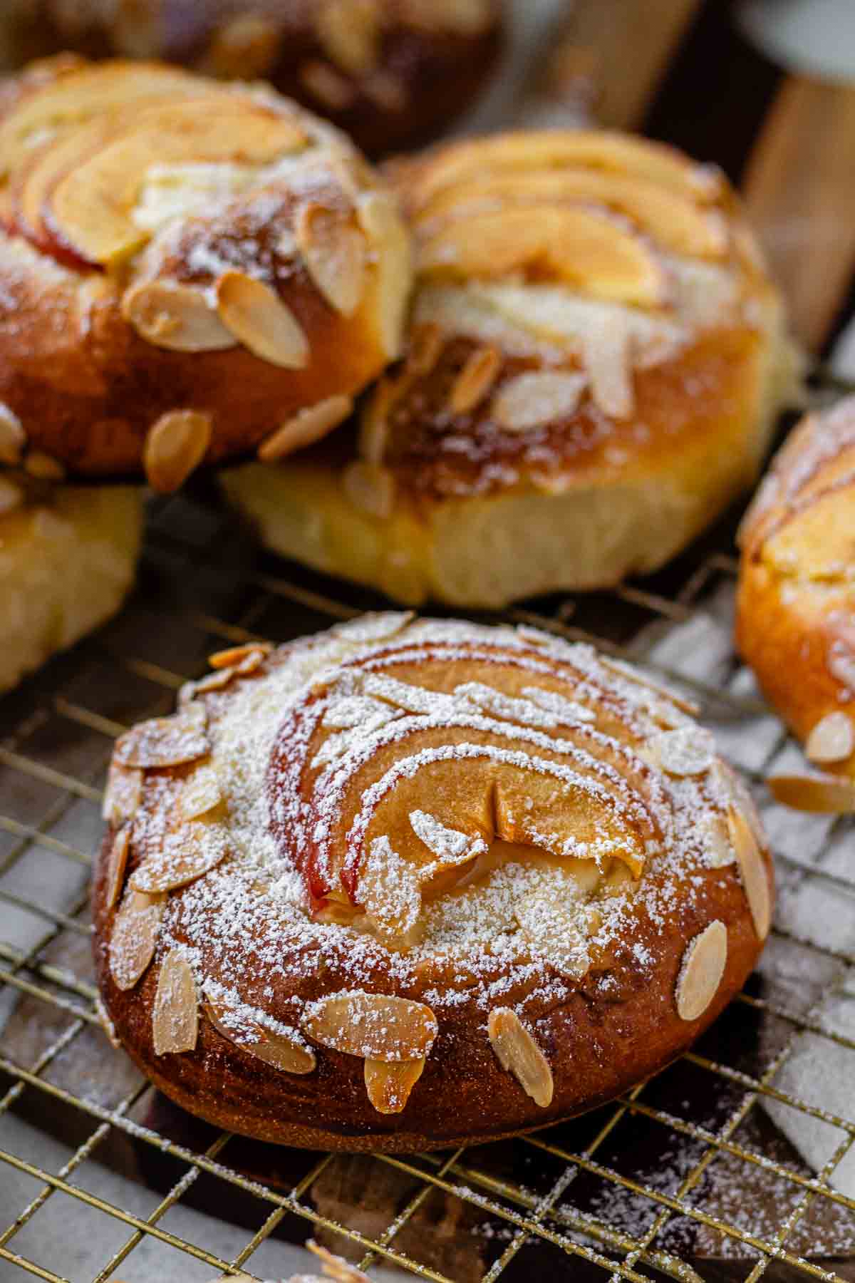 Apple and almond sweet brioche buns topped with sliced apples, cream cheese, powdered sugar, and toasted almonds on a baking sheet.