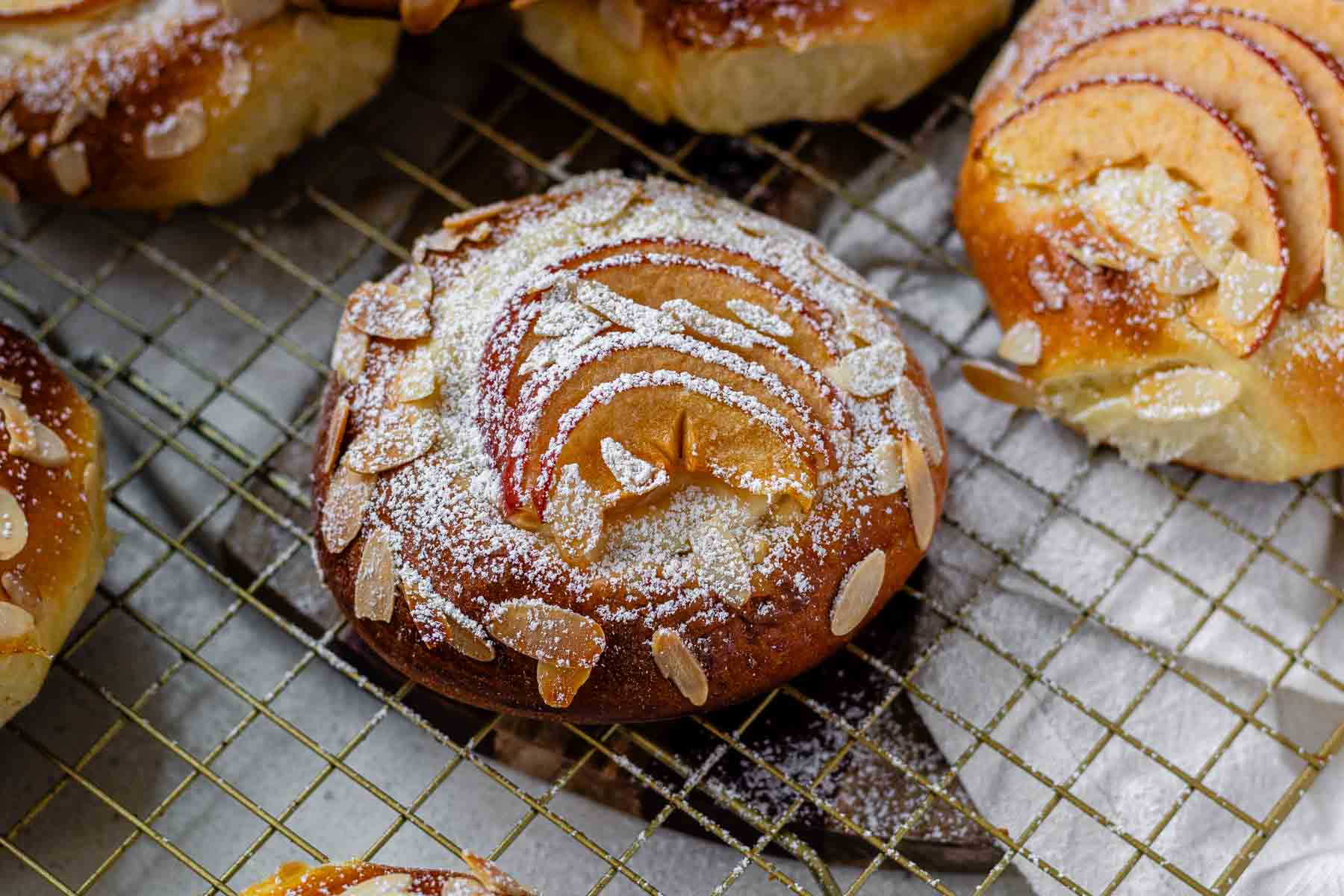Apple and almond sweet brioche buns topped with sliced apples, cream cheese, powdered sugar, and toasted almonds on a baking sheet.
