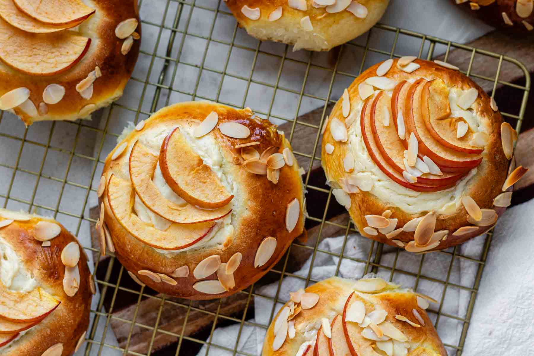 Apple and almond sweet brioche buns topped with sliced apples, cream cheese, and toasted almonds on a baking sheet.