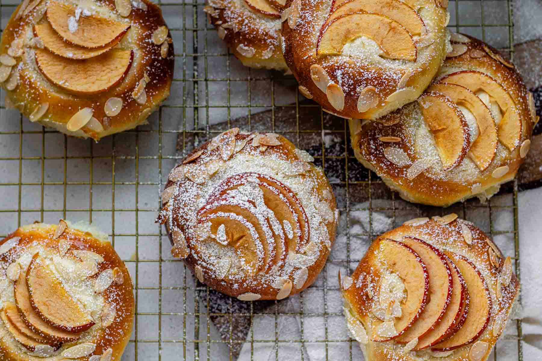Apple and almond sweet brioche buns topped with sliced apples, cream cheese, powdered sugar, and toasted almonds on a baking sheet.