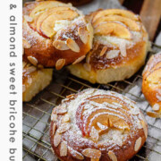 Apple and almond sweet brioche buns topped with sliced apples, cream cheese, powdered sugar, and toasted almonds on a baking sheet with the text apple & almond sweet brioche buns.