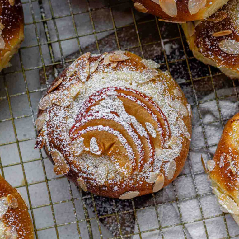 Apple and almond sweet brioche buns topped with sliced apples, cream cheese, powdered sugar, and toasted almonds on a baking sheet.