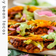Tacos on a platter with a green sauce and pickled red onions with the text crispy bean tacos.