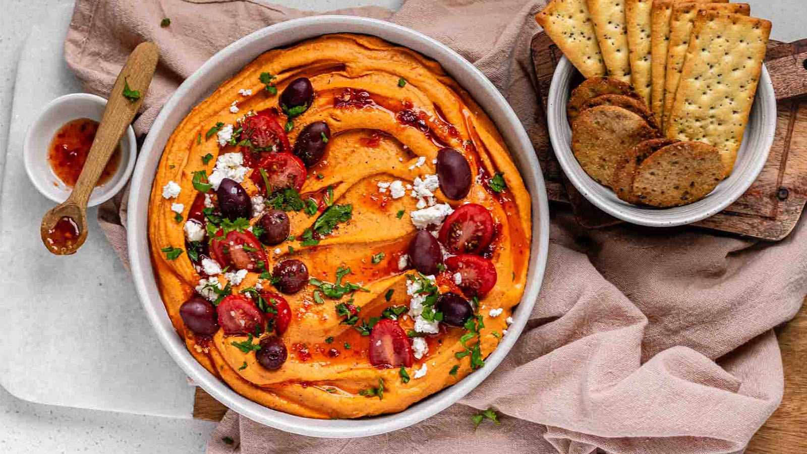 Orange hummus in a bowl garnished with olives, tomato, cheese, and chopped herbs.